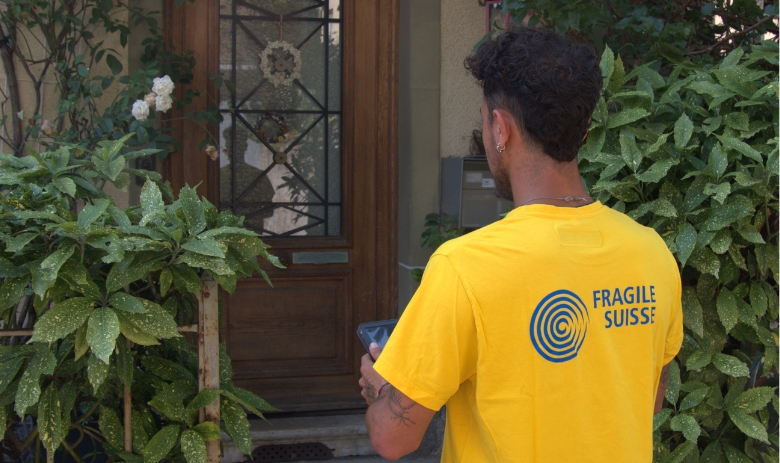 Dialoger von FRAGILE Suisse. Ein junger Mann mit gelbem FRAGILE Suisse-T-Shirt steht vor einer Haustüre mit einem Tablet in den Händen.