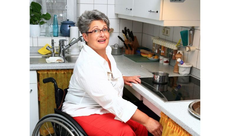 Vivre en toute autonomie Une femme aux cheveux courts gris et portant des lunettes est assise dans un fauteuil roulant devant la cuisinière dans sa cuisine.