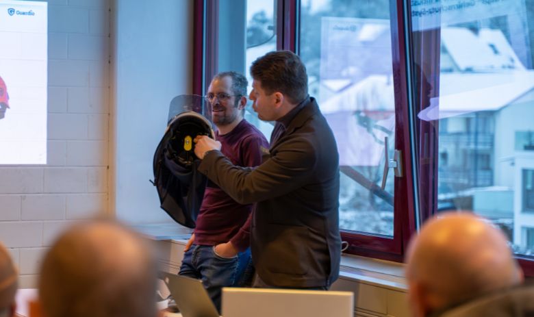 Prévention des accidents grâce au port du casque Deux hommes se tiennent devant un public. L'un des deux hommes montre un casque.
