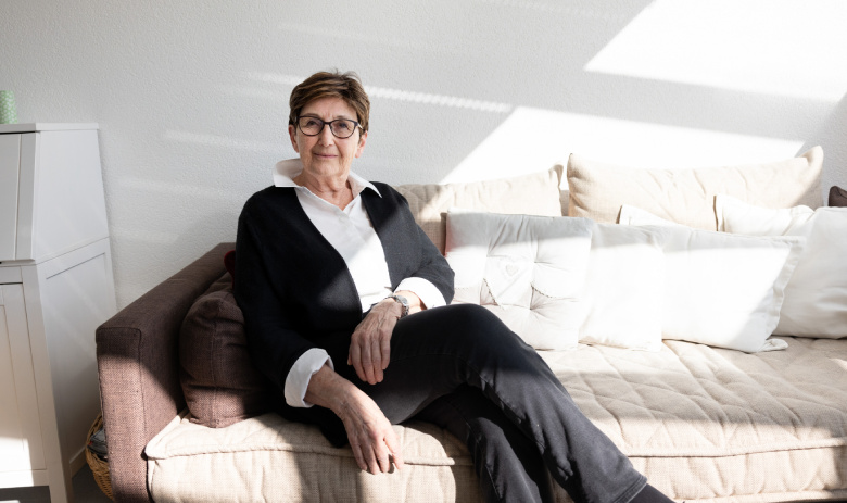 Josiane Parisod Josiane est assise sur un canapé. La pièce est très lumineuse, elle porte elle-même des vêtements sombres.
