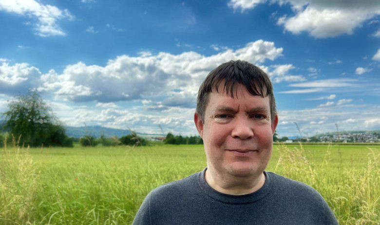 Peter F. Ein Mann blickt in die Kamera. Er steht draussen in der Natur. Im Hintergrund eine grüne wiese und ein tiefblauer Himmel mit weissen Wolken.