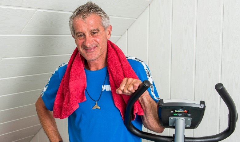 Daniel S. Portrait d'un homme debout à côté de son home-trainer. Il porte un t-shirt de sport bleu et un foulard rouge autour de la nuque.