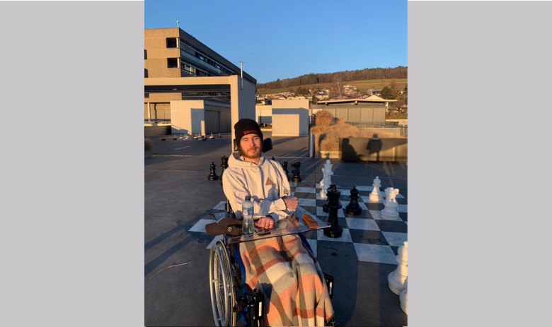 Un jeune homme est dans une chaise roulante, derrière lui se tient un échiquier géant